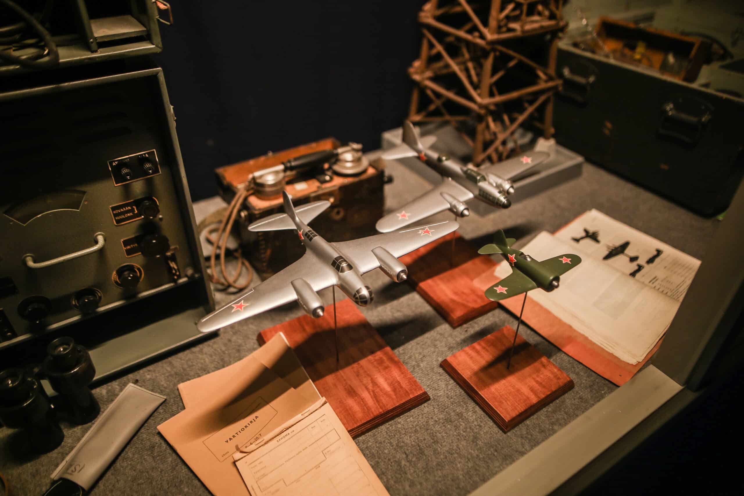 Kuva vitriinistä viestinäyttelyssä. Viholliskoneen pienoismalli, lentokoneen tunnistusopas ja muita käsikirjoja sekä radioita.